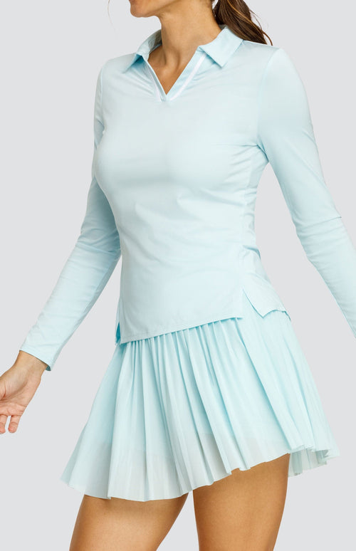 Model wearing a light blue long sleeve top with an open v neck foldover collar with white accents and side vents. Paired with a matching mesh pleated skort with solid undershorts.