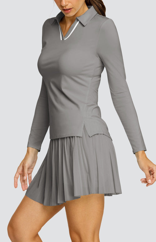 Model wearing a gray long sleeve top with an open v neck foldover collar with white accents and side vents. Paired with a matching mesh pleated skort with solid undershorts.