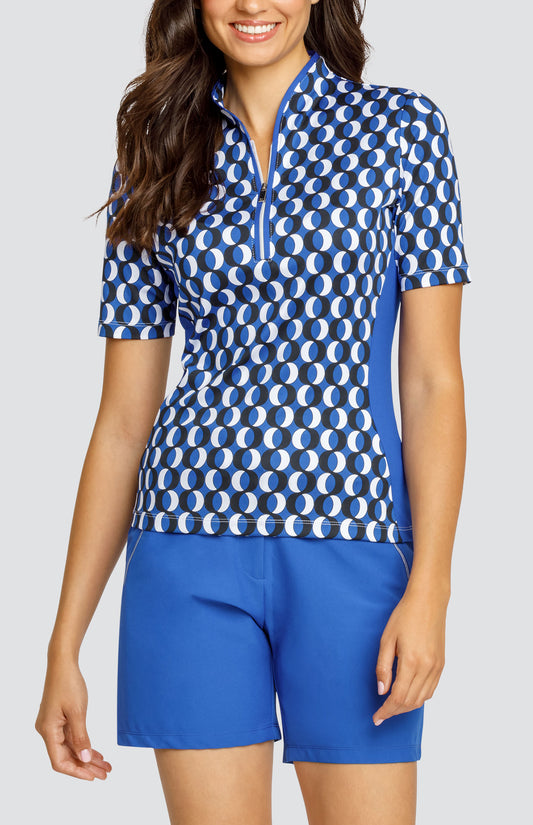 Model wearing a short sleeve golf top with a circular blue, white, and black pattern, and blue shorts.