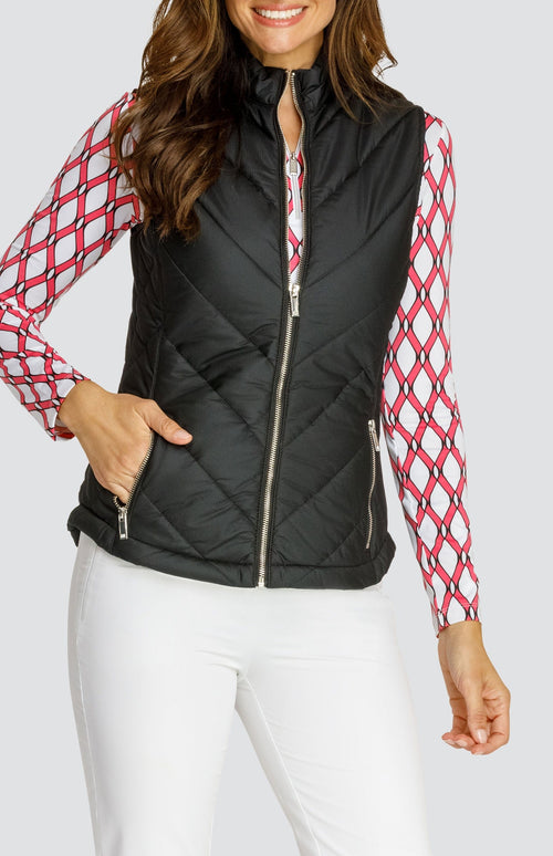 Model wearing a black puffer vest over a long sleeve quarter-zip golf top with a pink and white lattice pattern and a pull on pink golf skort with a flounce insert.