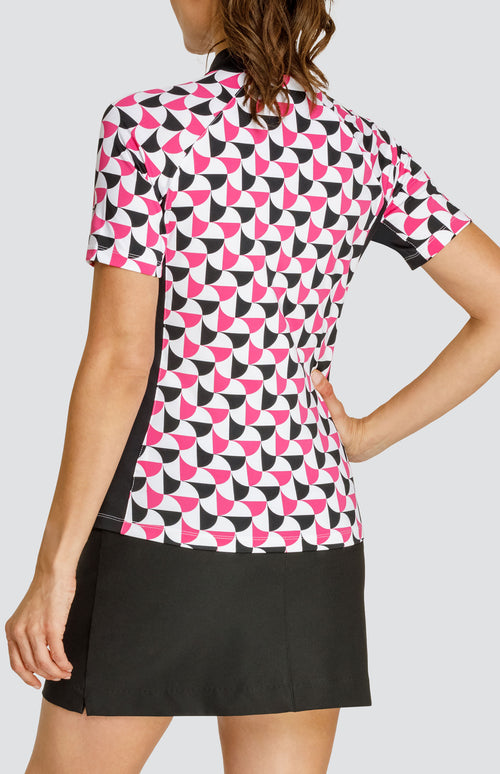 Model wearing a short sleeve quarter-zip golf top in a pink and black alternating triangular print on a white background with black side and underarm inserts, paired with a solid black pull-on golf skort.