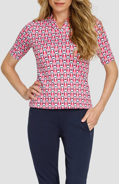 Front view of a woman wearing a mid-length sleeve golf top with a star collar neckline. The print is a geometric pattern of 6-sided gemstones in a light pink color on a white background. She is also wearing dark navy blue pants with zipper pockets.