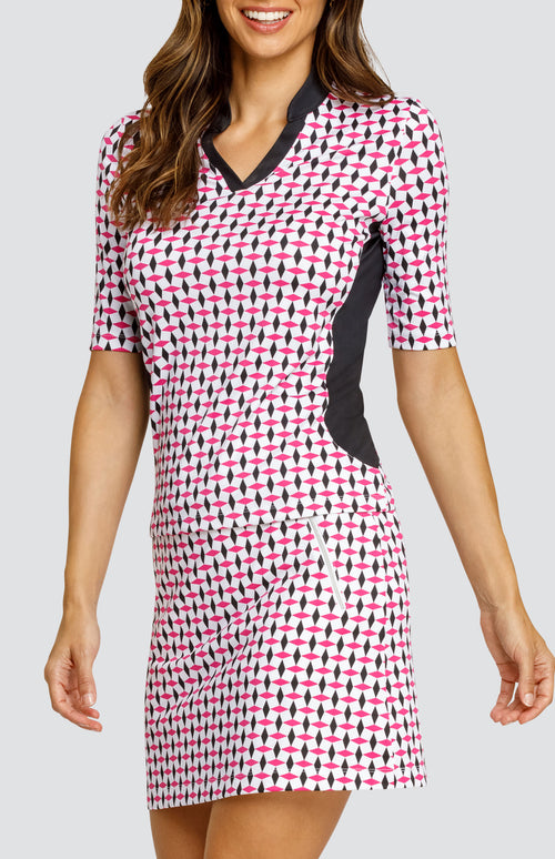 Model wearing an open neck elbow sleeve golf top in a pink and black geometric alternating diamond print on a white background with black side inserts, paired with a pull-on skort in a matching print.