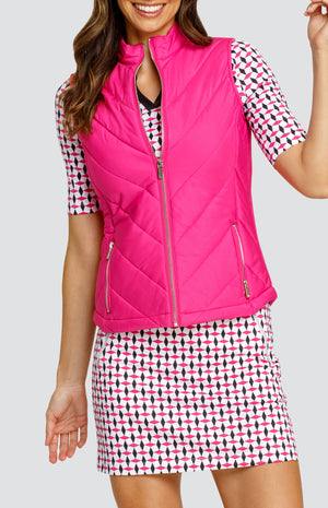 Model wearing a solid pink zip-up puffer vest, paired with an open neck elbow sleeve golf top in a pink and black geometric alternating diamond print on a white background with black side inserts and a pull-on skort in a matching print.