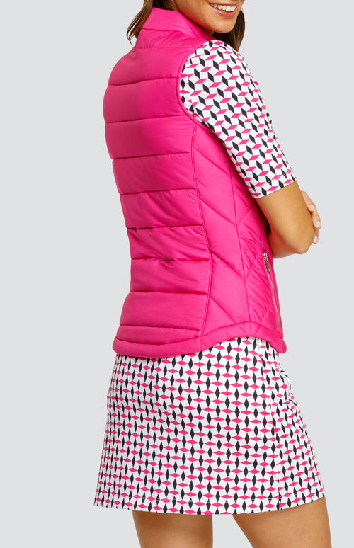 Model wearing a solid pink zip-up puffer vest, paired with an open neck elbow sleeve golf top in a pink and black geometric alternating diamond print on a white background with black side inserts and a pull-on skort in a matching print.