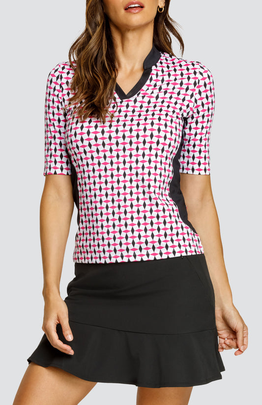 Model wearing an open neck elbow sleeve golf top in a pink and black geometric alternating diamond print on a white background with black side inserts, paired with a solid black pull-on skort.