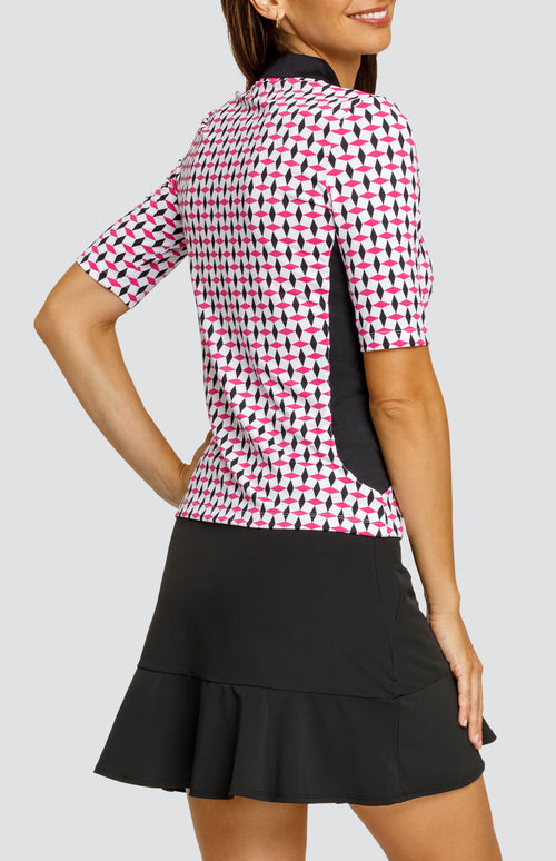 Model wearing an open neck elbow sleeve golf top in a pink and black geometric alternating diamond print on a white background with black side inserts, paired with a solid black pull-on skort.