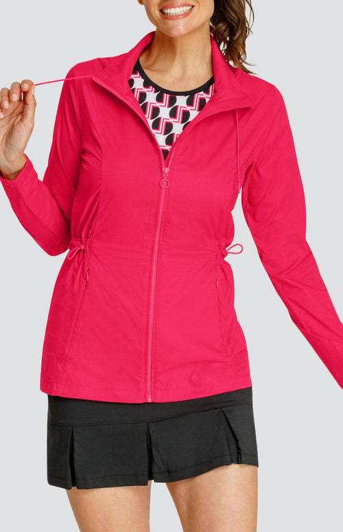 Model wearing a pinkish red water resistant jacket with a drawstring waist and attached hood, over a tank with geometric pattern and black pleated skort.